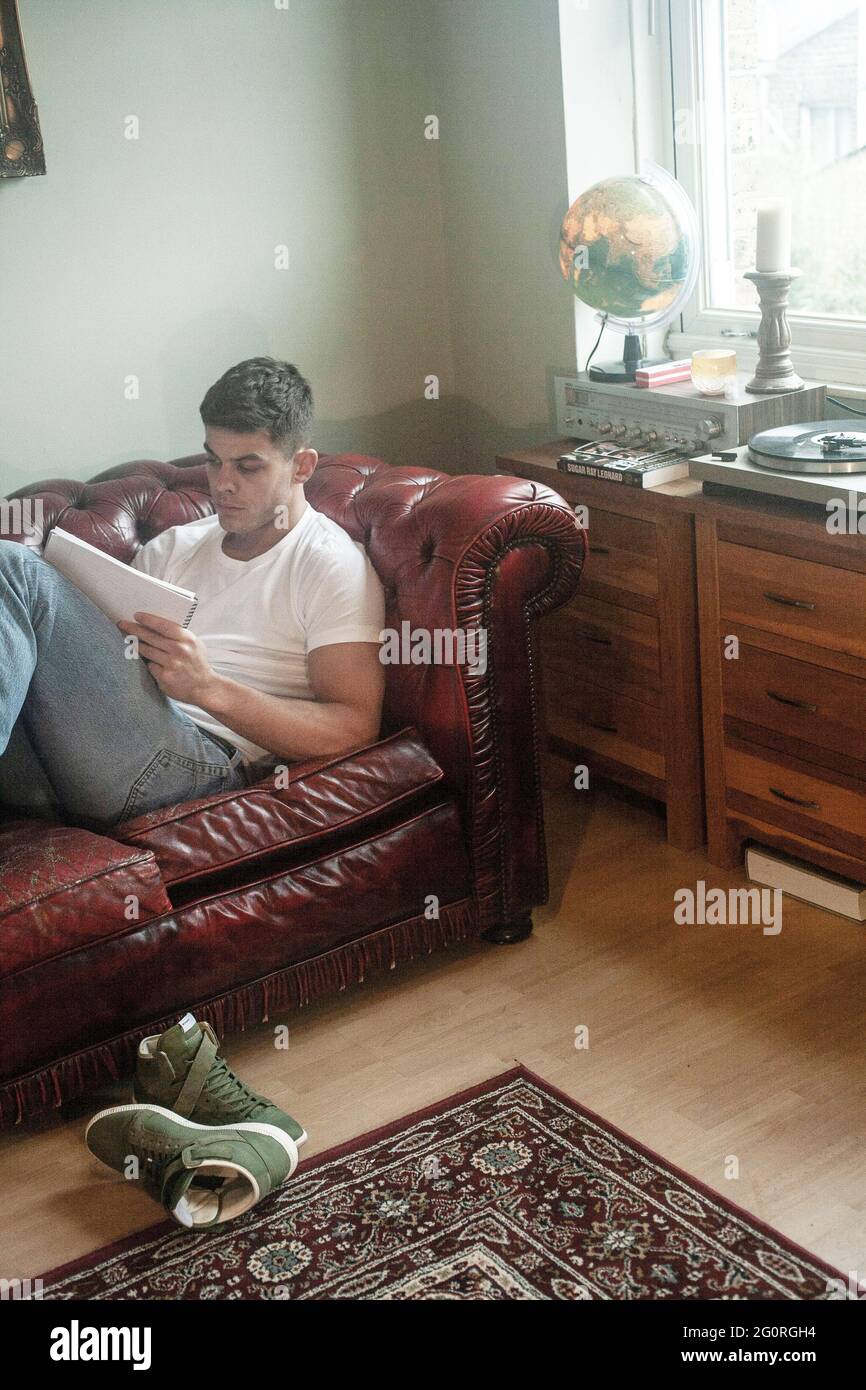 A young man is sitting on a sofa and writing a letter in room. Stock Photo