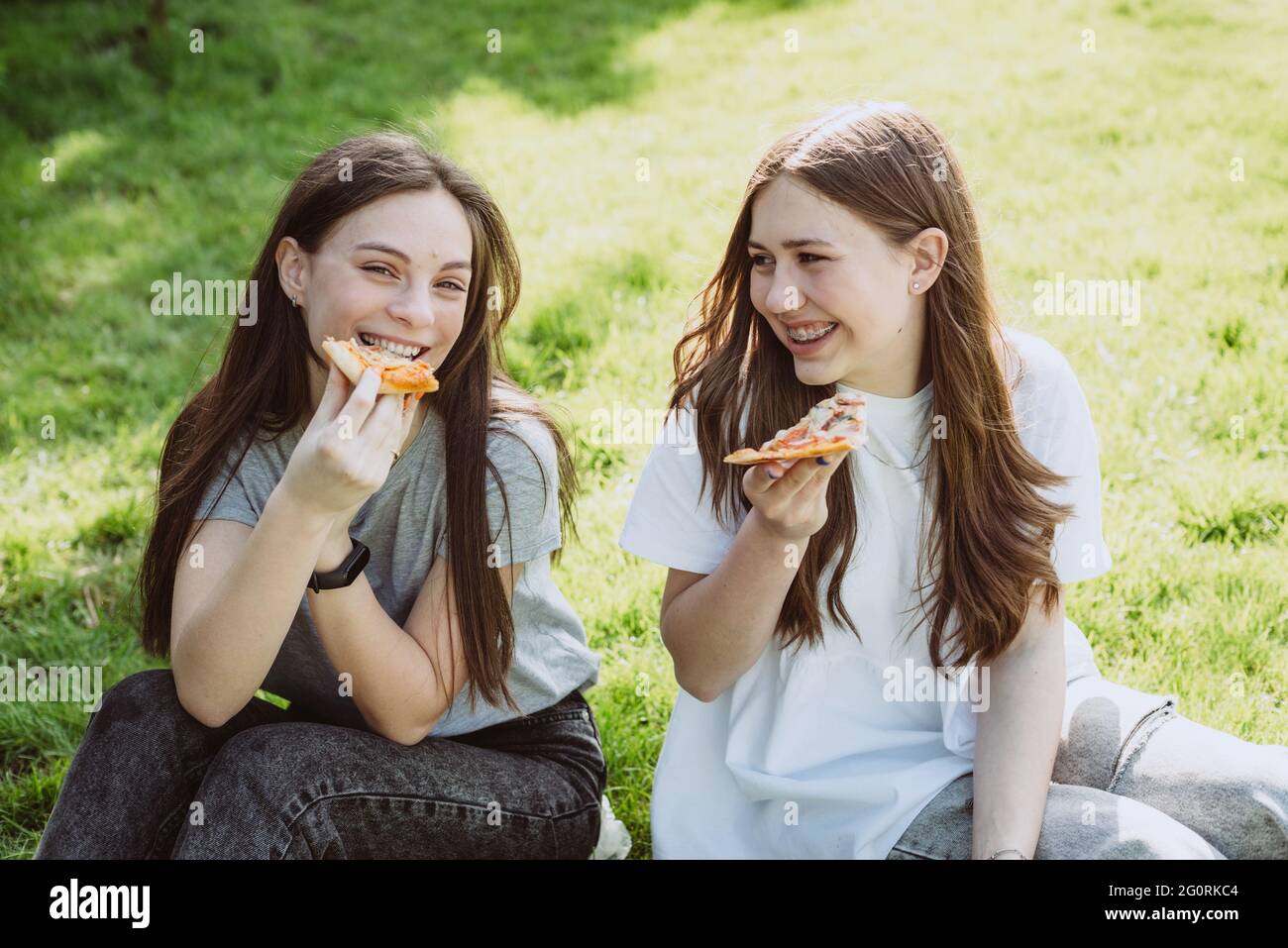 Two cheerful young teen friends in the park eating pizza. Women eat fast food. Not a healthy diet. Soft selective focus. Stock Photo