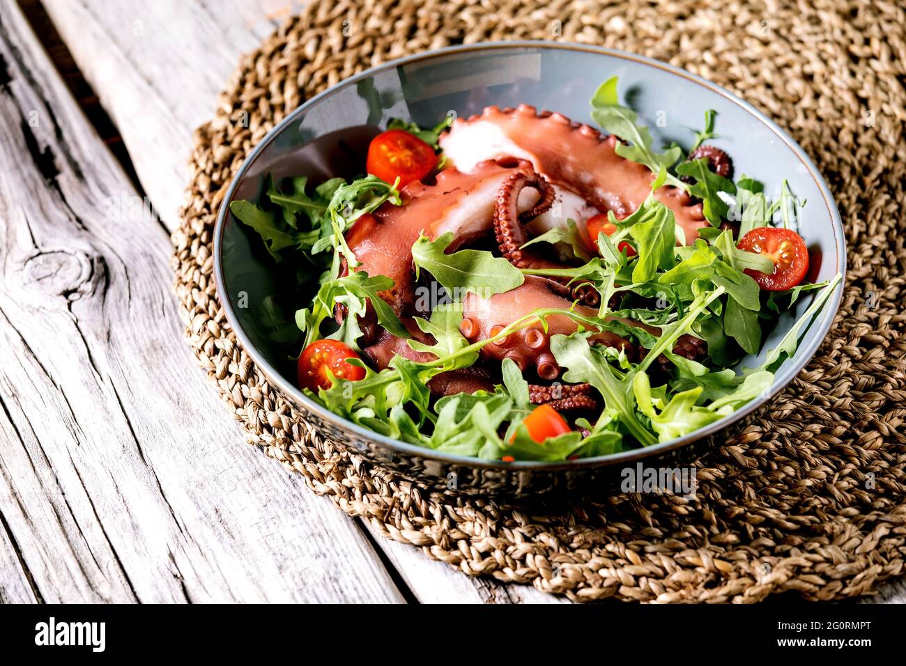 Seafood salad. Coocked tentacles of octopus on blue ceramic plate served with rocket leaf aragula and cherry tomato salad over grey wooden background Stock Photo