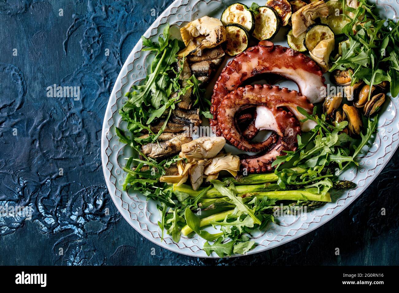 Seafood salad. Coocked grilled tentacles of octopus, sardines and mussels on blue ceramic plate with arugula salad, zucchini and asparagus over blue t Stock Photo