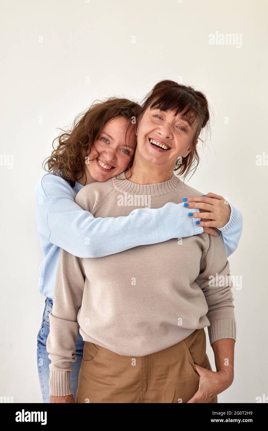 Young woman hugs happy mature smiling lady wearing casual clothing stand together in apartment room with beige wall closeup Stock Photo