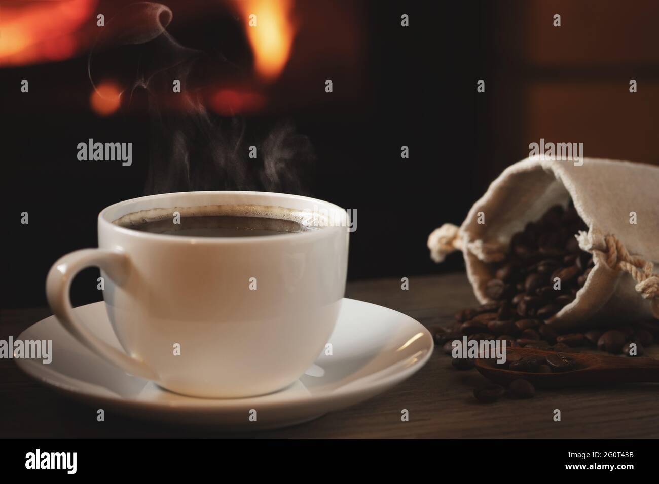 Close-up of cup of hot coffee and coffee beans in a bag on a wooden table in front of a burning fireplace. Selective focus. Stock Photo
