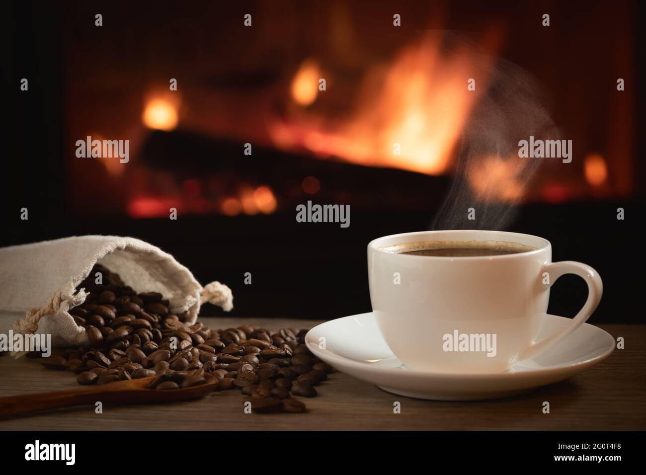 Cup of hot coffee and coffee beans in a bag on a wooden table in front of a burning fireplace. Selective focus. Stock Photo