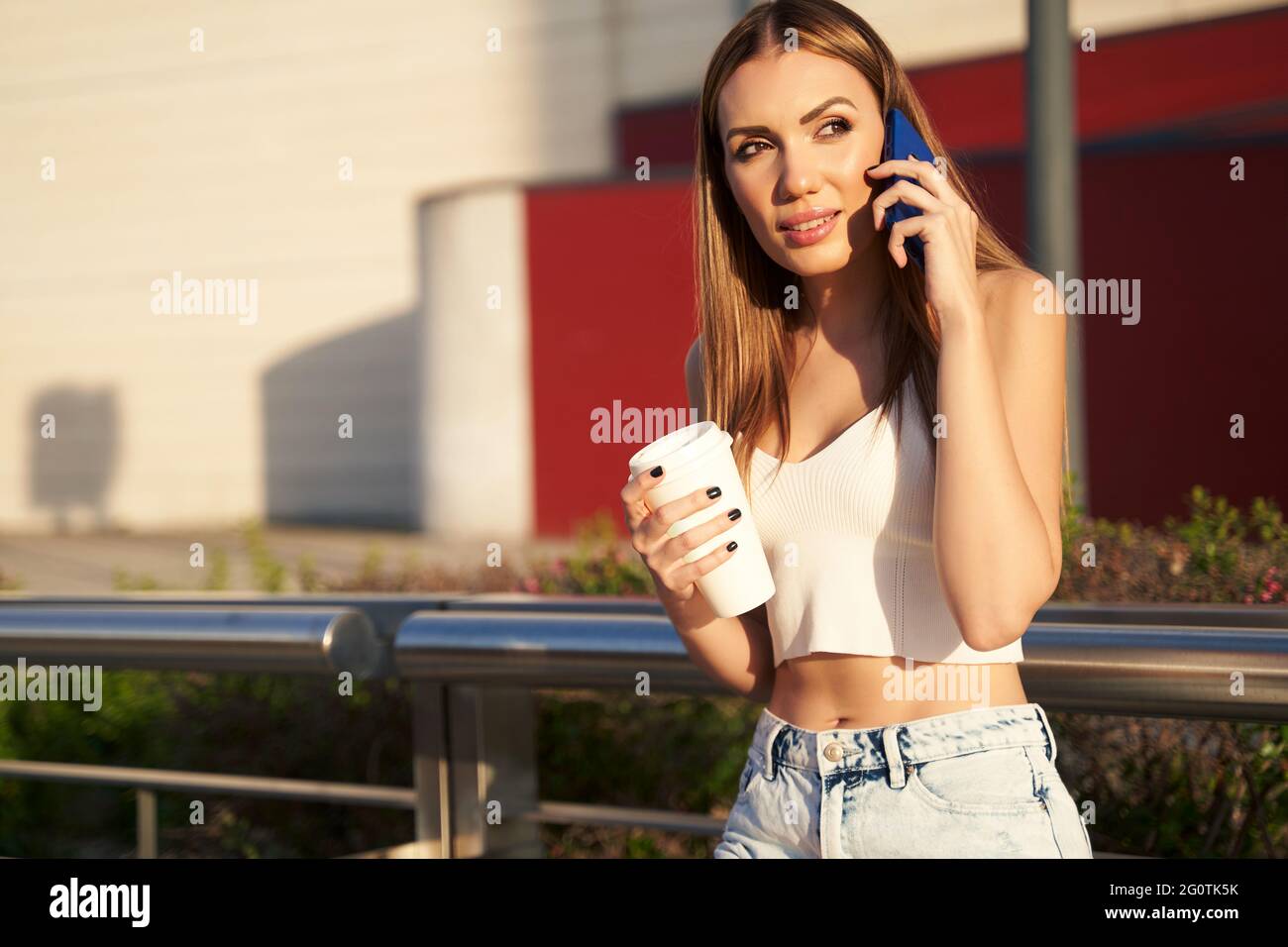 Young woman waiting for city train and talking on phone with coffee in hand. Passenger at streetcar stop calling with smartphone. Concept of public transport and communication. High quality photo Stock Photo