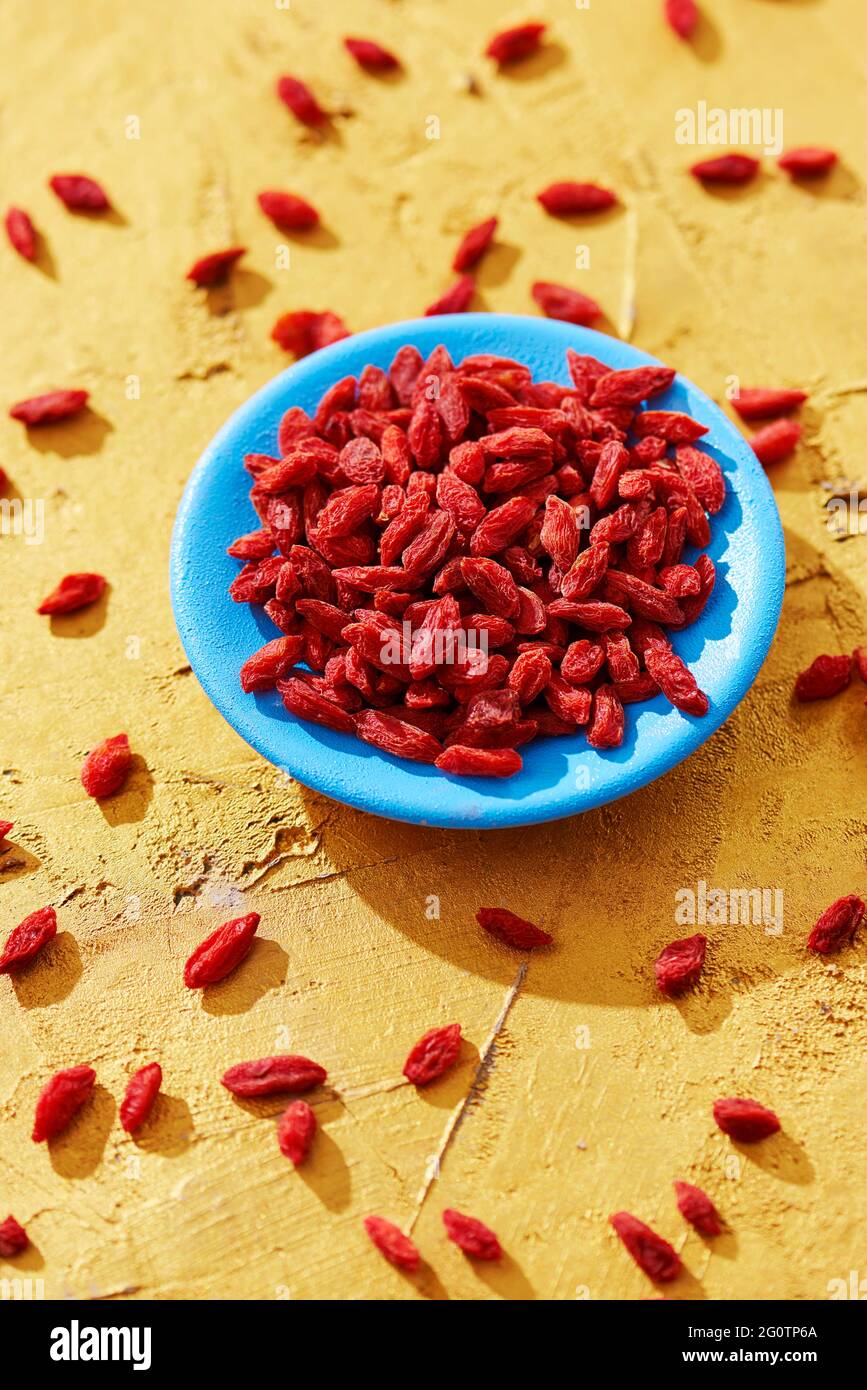 closeup of some dried goji berries in a blue plate and some other berries sprinkled on a golden textured surface Stock Photo