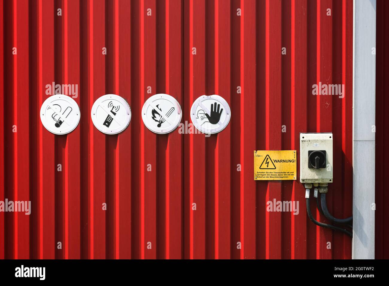 Red wall made of metal profiles with four round warning signs,   a circuit breaker and a rectangular yellow warning label Stock Photo