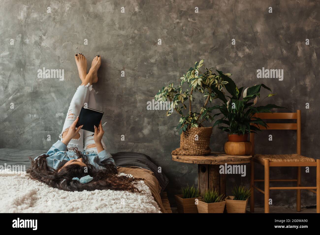Woman lies on bed with her legs crossed up, with book in her hands. Stock Photo