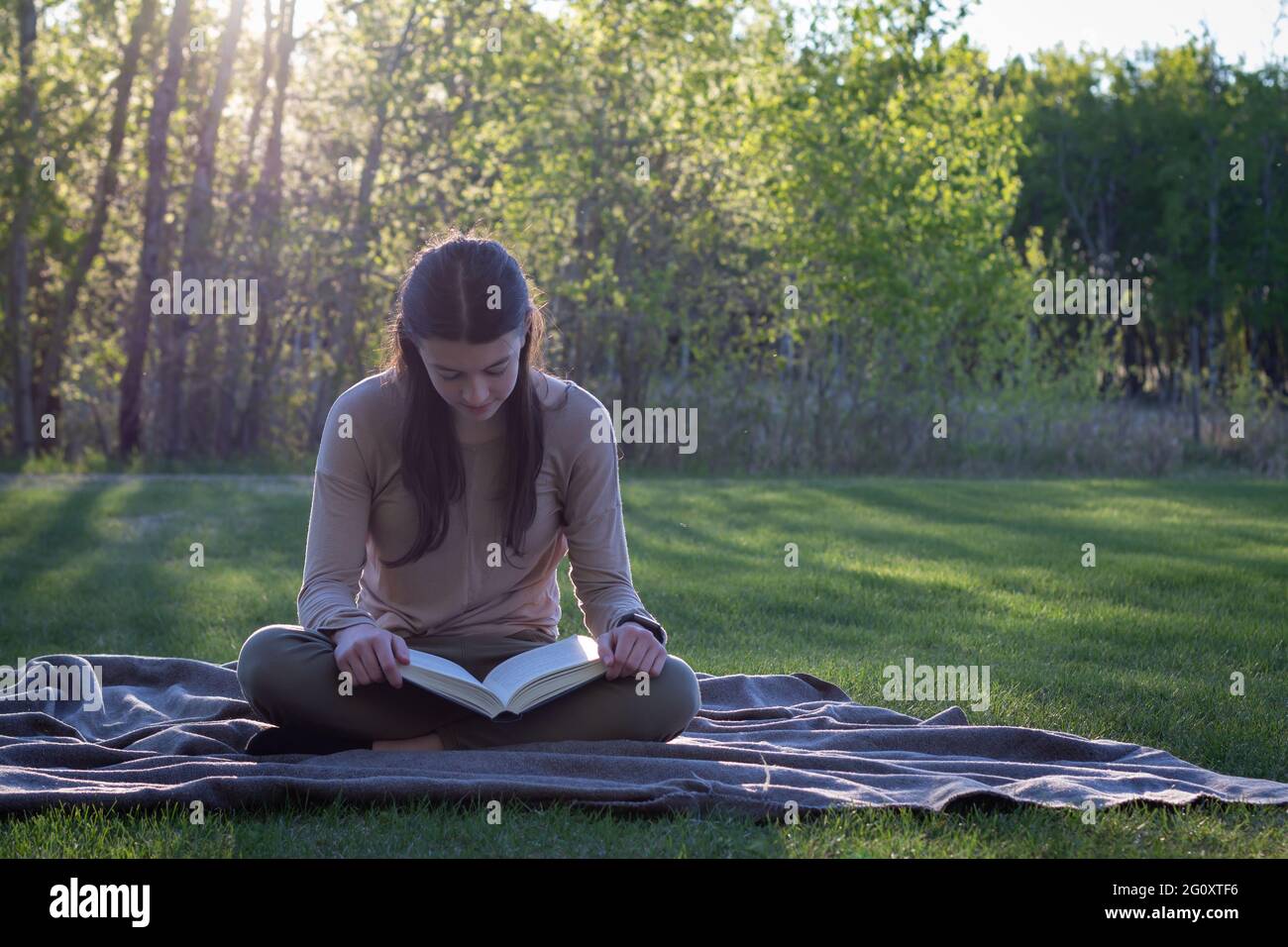young woman reading book while sitting on a blanket outdoors in the summer with beautiful landscape, green grass and trees Stock Photo