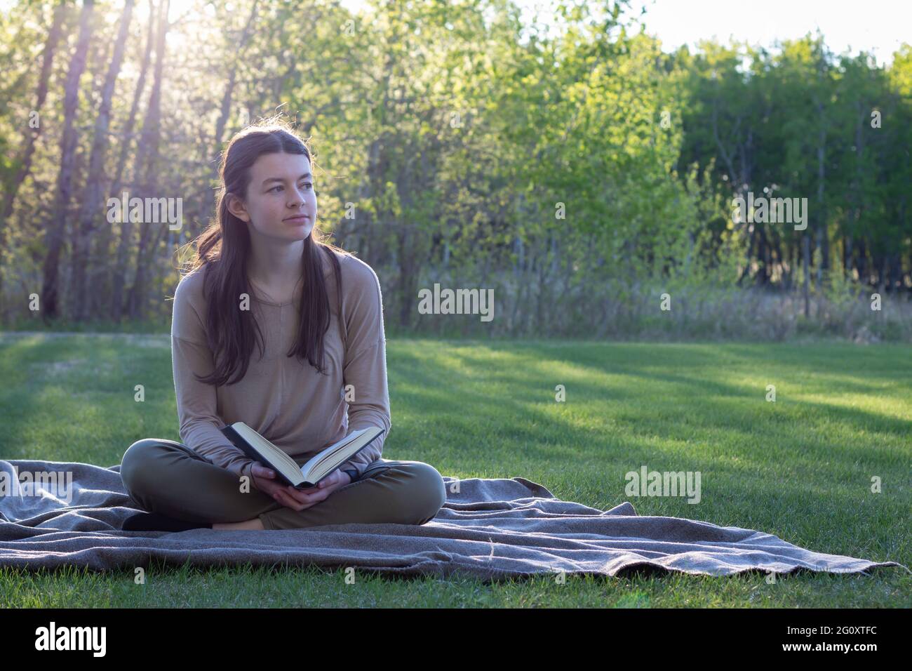 young woman reading book while sitting on a blanket outdoors in the summer with beautiful landscape, green grass and trees Stock Photo