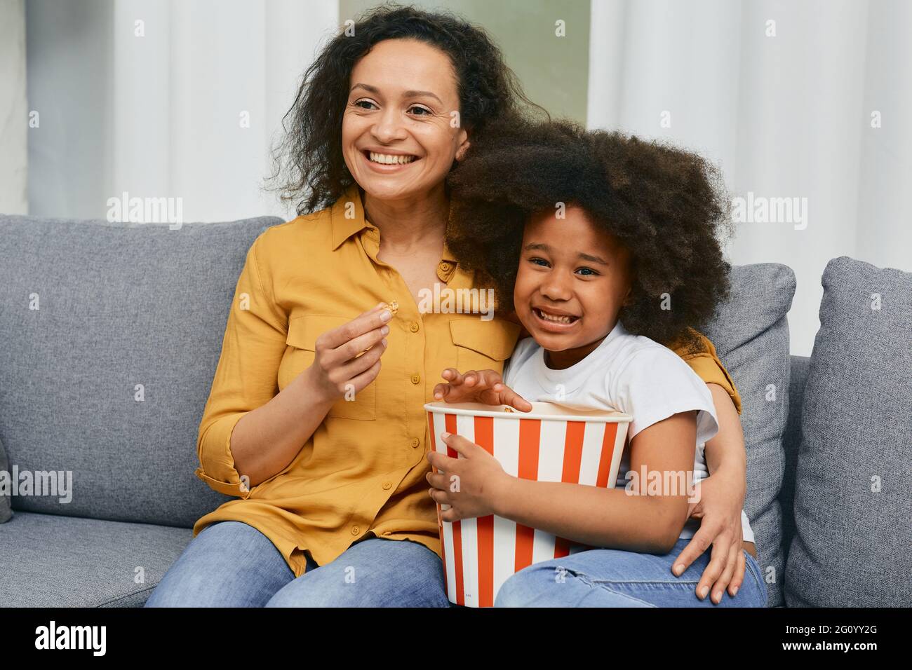 African American mother with her little daughter hugging and watching cinema together and eating popcorn from a large striped bucket Stock Photo
