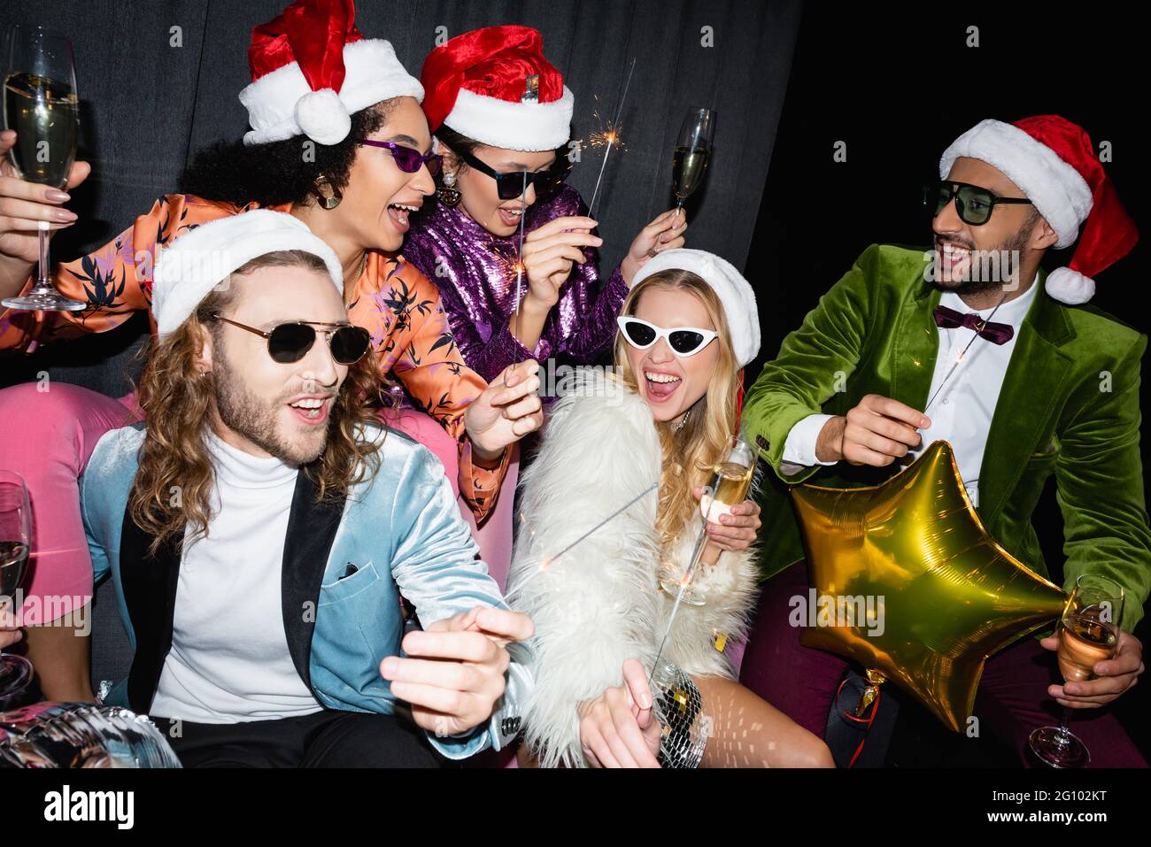 interracial friends in santa hats drinking champagne and celebrating new year near grey curtain on black background Stock Photo