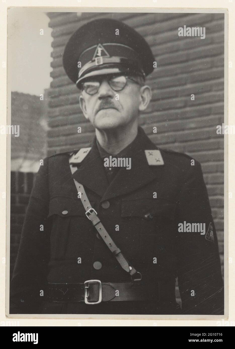 Portrait of a member of the WA; NSB. Portrait of a member of the WA. Photographed from the waist, standing in front of a house. On his collar a distinctive with two stars: companion, the lower frame. Hat with the Wolfsangel, on his left sleeve. The triangular emblem of the NSB. Belt diagonally over the chest. It is an older man with glasses and mustache. This concerns the same man as at NG-2007-35-206, so it would be a member of the country guard, founded at the end of 1943. Stock Photo
