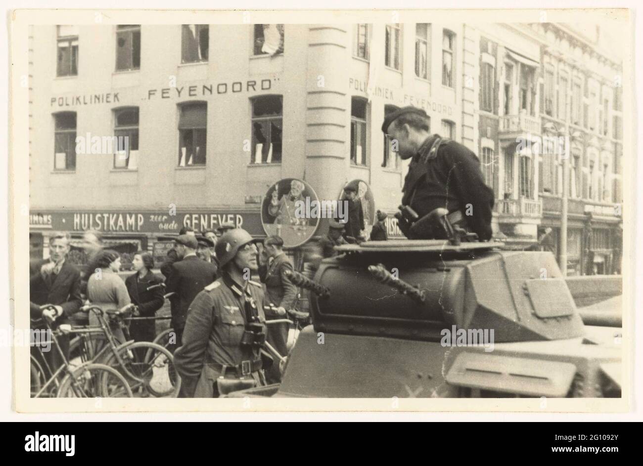 German army in Rotterdam. German Wehrmacht soldiers, one in a tank with a cap and binoculars, talk to each other on the street in Rotterdam South. The one left has a helmet, wears glasses and binoculars around his neck. The Feyenoord clinic can be seen in the background. To the right are Dutch, with cycling, talking. It would be German tanks of the 9th Armored Division that make contact with the men's men's men's men. The photo is probably made around the capitulation of the Netherlands on May 15, 1940. Stock Photo