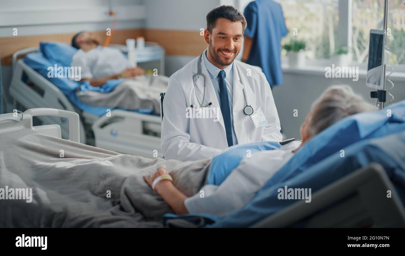 Hospital Ward: Friendly Doctor Talks to Beautiful Senior Female Patient Resting in Bed, Explains Test Results, Gives Recovery Advice. Physician Talks Stock Photo