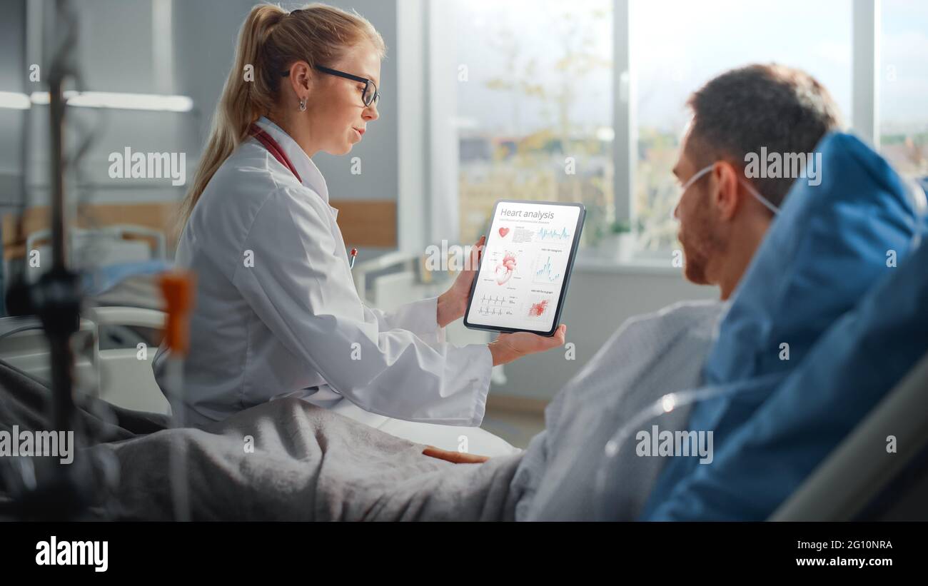 Hospital Ward: Friendly Female Doctor Talks to Sick Male Patient Resting in Bed, Uses Tablet Computer, Shows and Explains Heart Scan Results Stock Photo