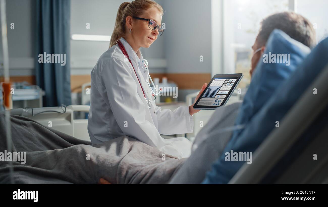 Hospital Ward: Friendly Female Doctor Talks to Sick Male Patient Resting in Bed, Uses Tablet Computer, Shows and Explains Lung Scan Results. Physician Stock Photo