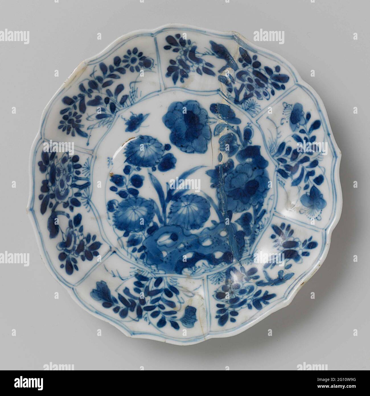 Saucer with Rocks, Flower Sprays and Butterflies. Dish of porcelain with ribbed wall and scalloped edge, painted in underglaze blue. On the flat a rock with two flower branches (chrysanthemum, peony) and a butterfly; the wall divided into six compartments filled with flower branches and birds; Three flower branches on the back. Marked on the underside with a seal mark in a double circle. Dish has been broken. Blue White. Stock Photo