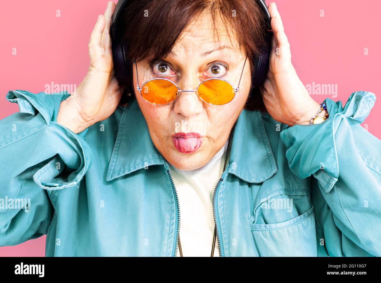 Portrait of middle aged woman listening to music with headphones - Close up of an older lady with orange hippie glasses on purple background - focus o Stock Photo