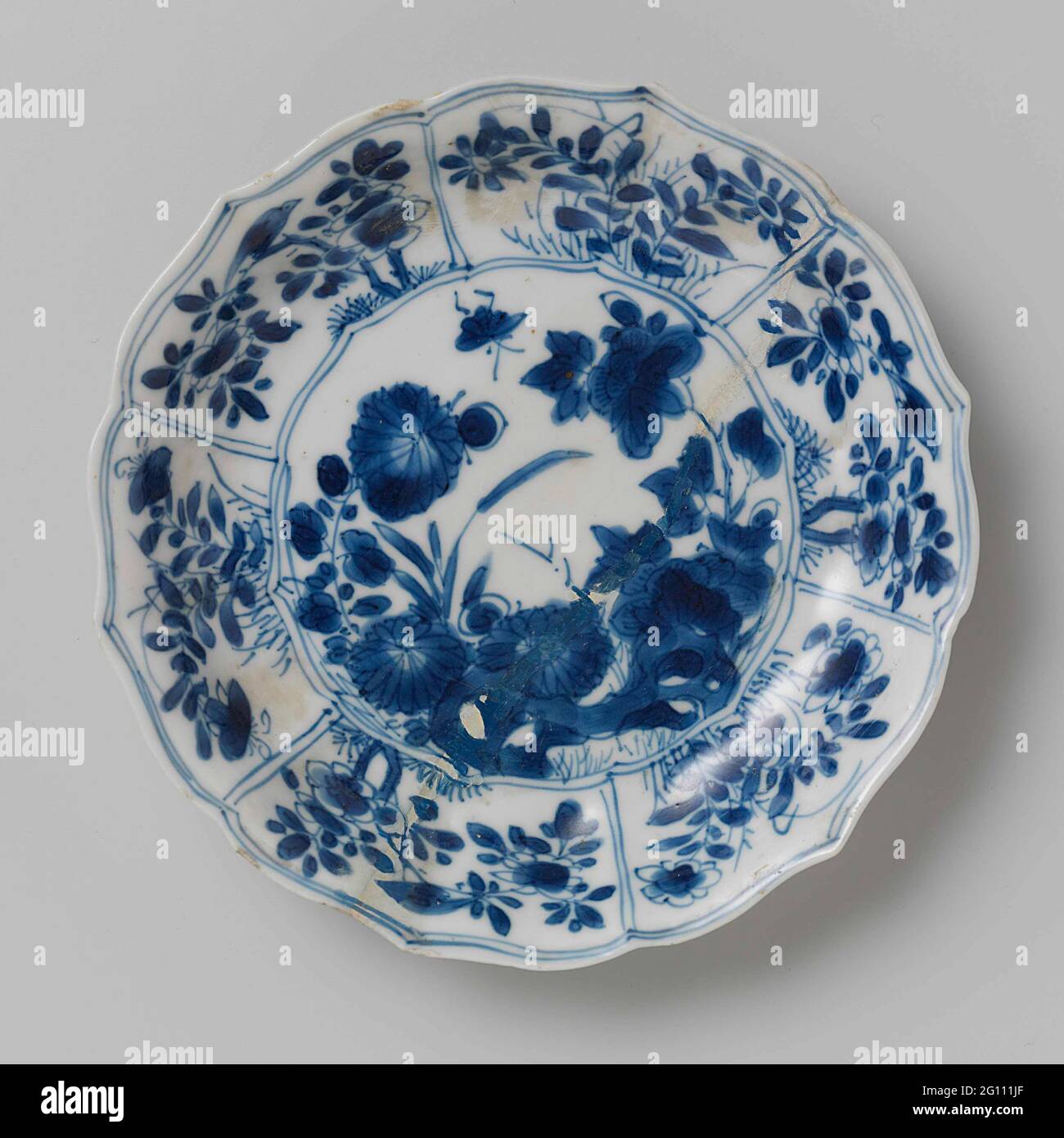 Saucer with Rocks, Flower Sprays and Butterflies. Dish of porcelain with ribbed wall and scalloped edge, painted in underglaze blue. On the flat a rock with two flower branches (chrysanthemum, peony) and a butterfly; the wall divided into six compartments filled with flower branches and birds; Three flower branches on the back. Marked on the underside with a seal mark in a double circle. Dish has been broken. Blue White. Stock Photo