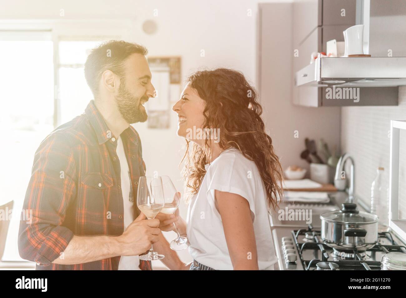 Young couple drinks a glass of white wine in the kitchen - couple in love celebrating their anniversary enjoying their new home - warm flare on backgr Stock Photo