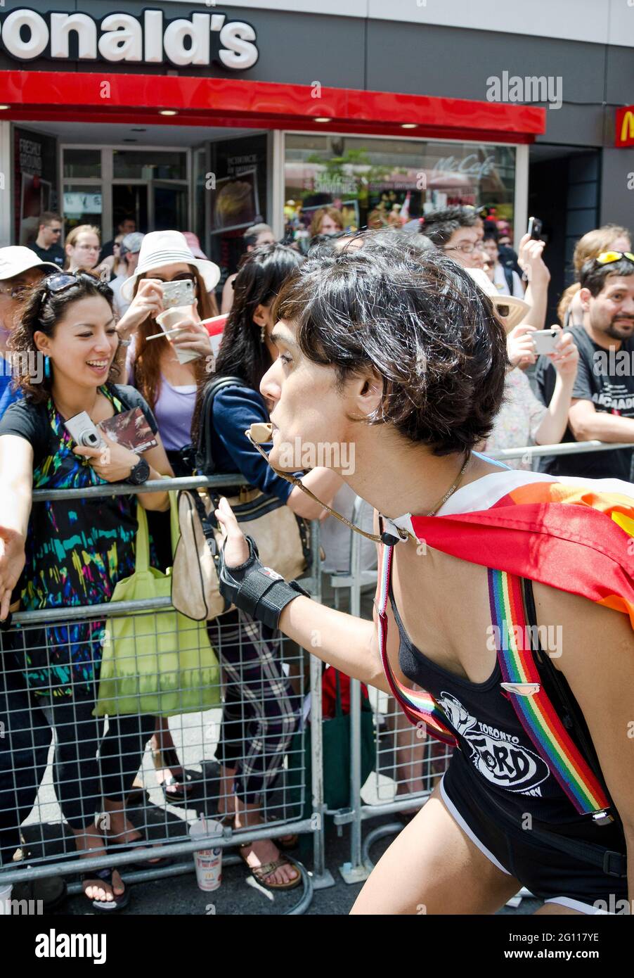 Woman blowing whistle at pride parade Toronto, wearing a cape and performing while passing in front of a crowd cheering from the sidelines Stock Photo