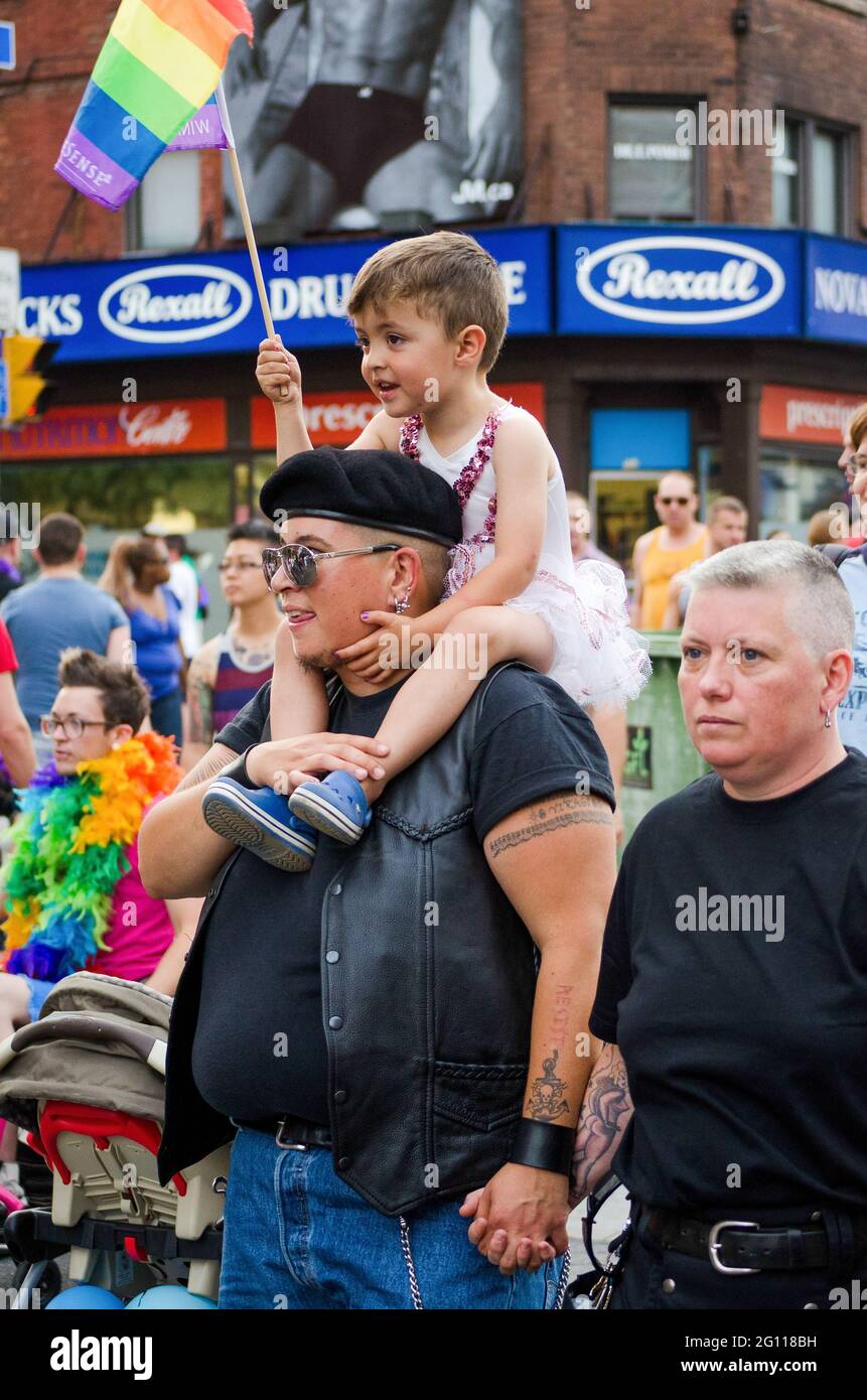 Couple with child on shoulder at pride parade in Toronto, child is wearing a white tutu and waving a rainbow flag against a backdrop of building, sign Stock Photo
