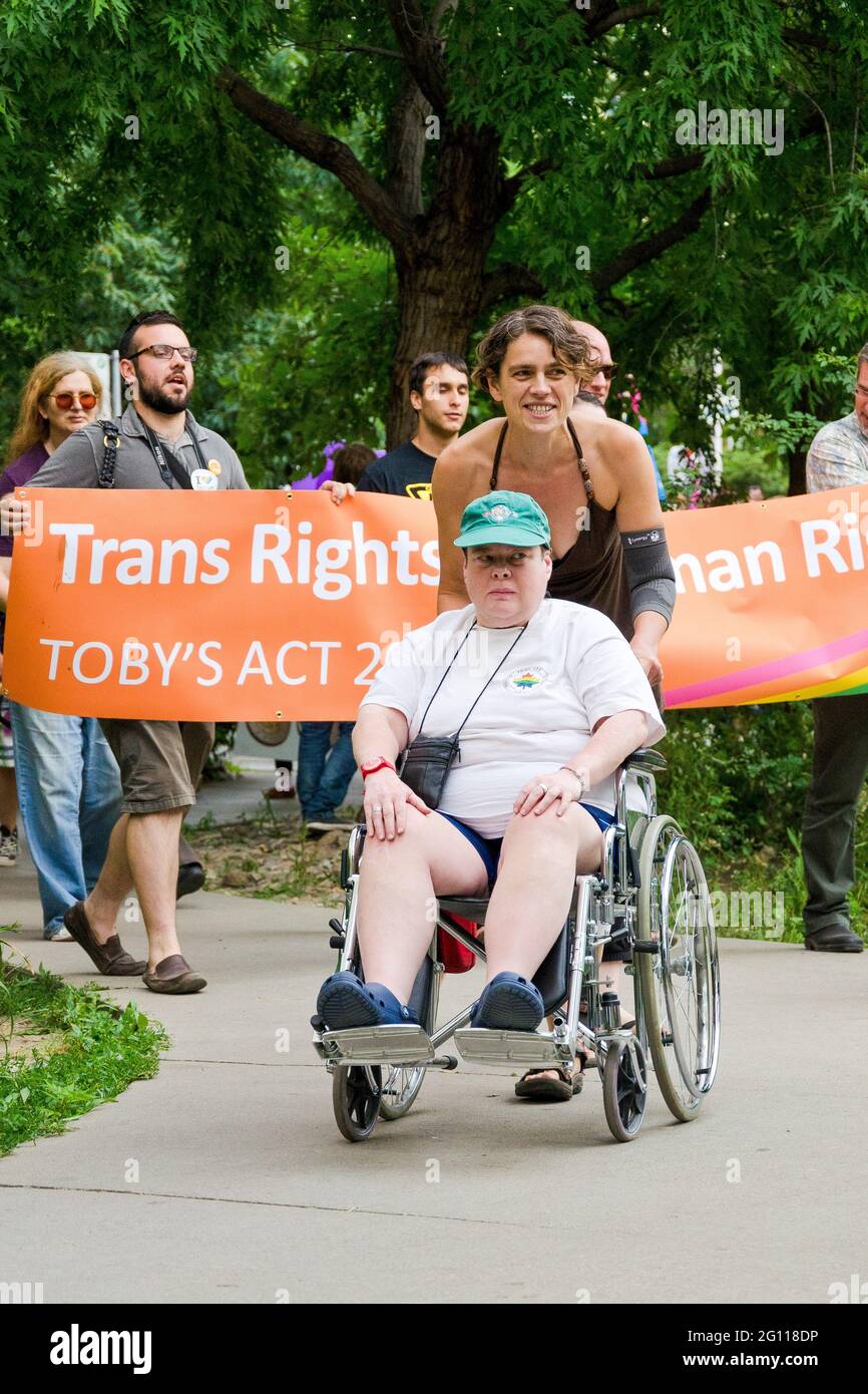 Wheelchair enabled participant in pride parade Toronto, wearing green baseball cap assisted by a woman dressed in a black halter dress, passing ahead Stock Photo