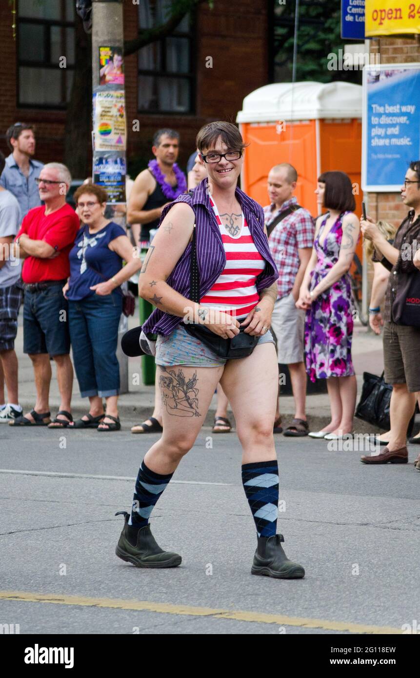Smiling woman at pride parade Toronto, full shot of a tattoed reveler wearing shorts and glasses walking down the street with out of focus spectators Stock Photo
