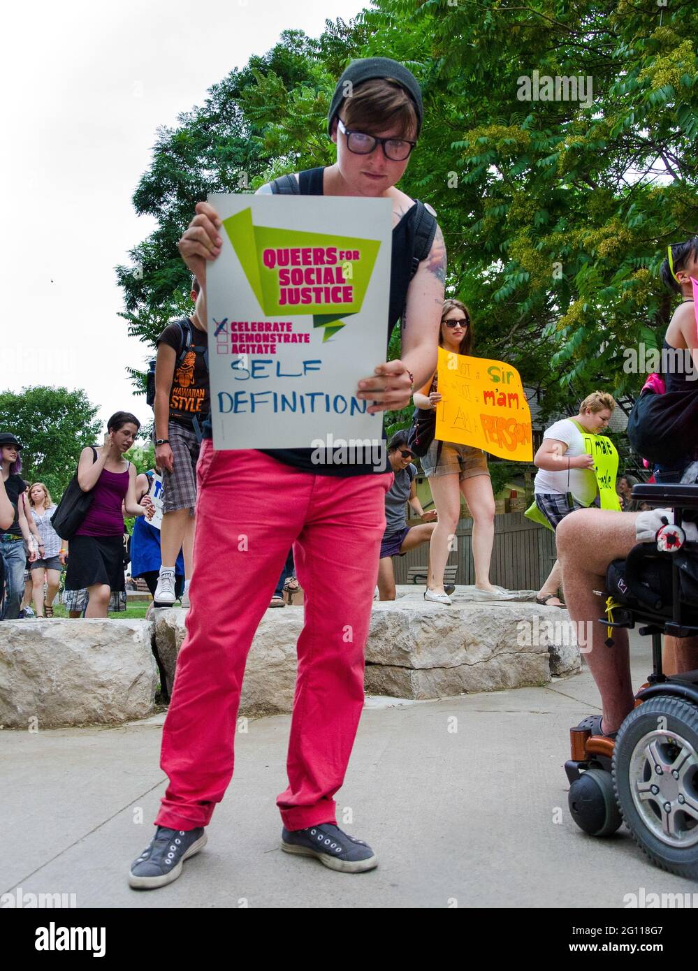Participant holding banner at pride parade Toronto, full shot of bespectacled woman standing on a garden path with more people closing in, in the back Stock Photo