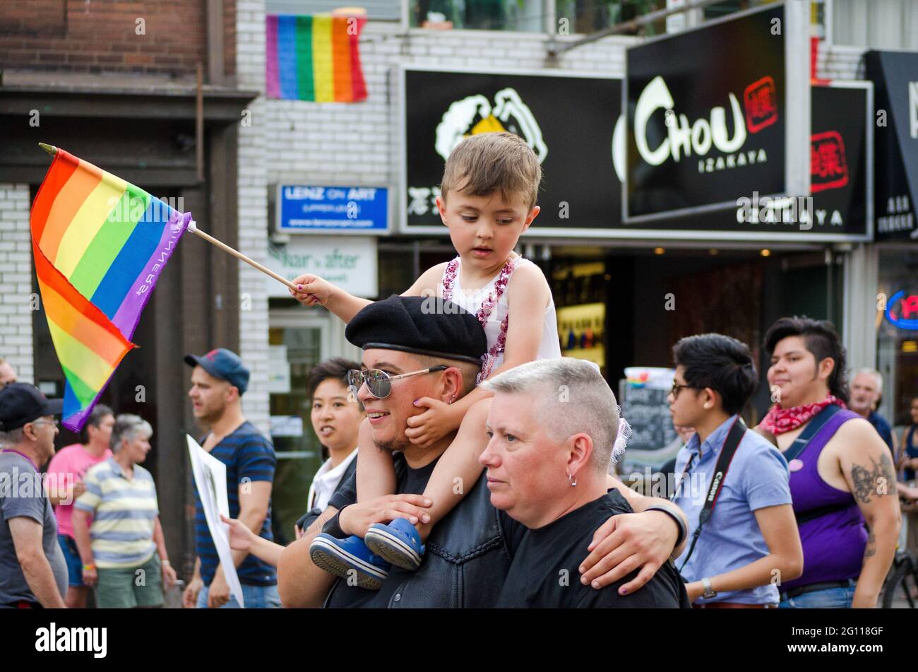 Couple with a child on shoulder at pride parade in Toronto, their closeup waving flag amidst a crowd of marchers in the background Stock Photo