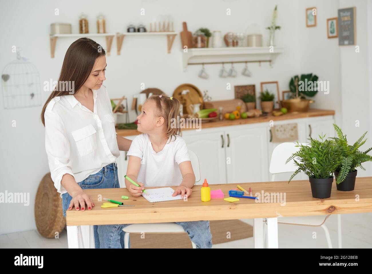 Tutor and child doing homework at kitchen. Mom and daughter are trying to solve the task. They are in a good mood and smile. Stock Photo