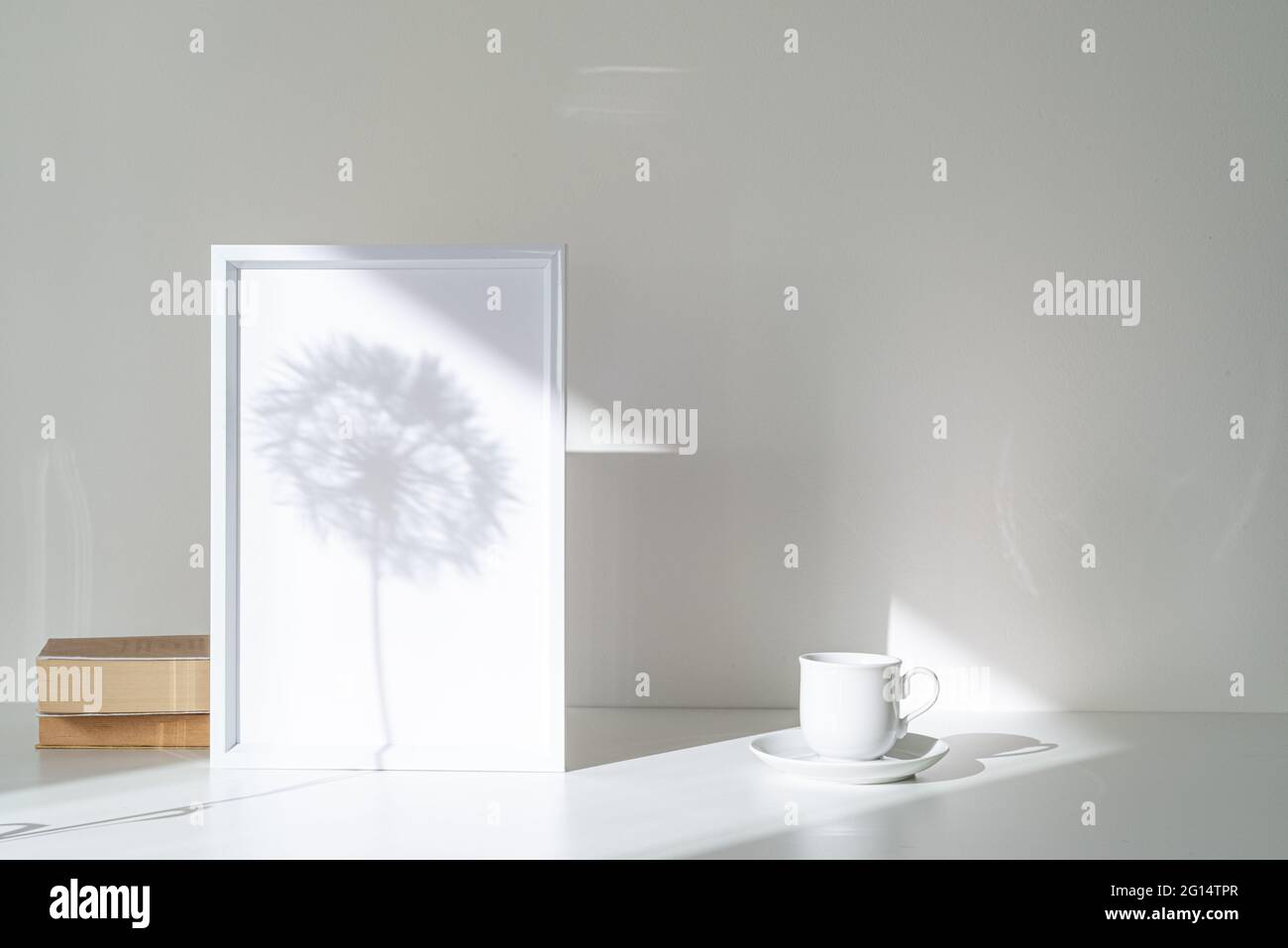 Empty white picture frame mockup in sunlight. Long shadow effect of shower head in the frame. Books and cup of coffee. Elegant lifestyle. White table Stock Photo