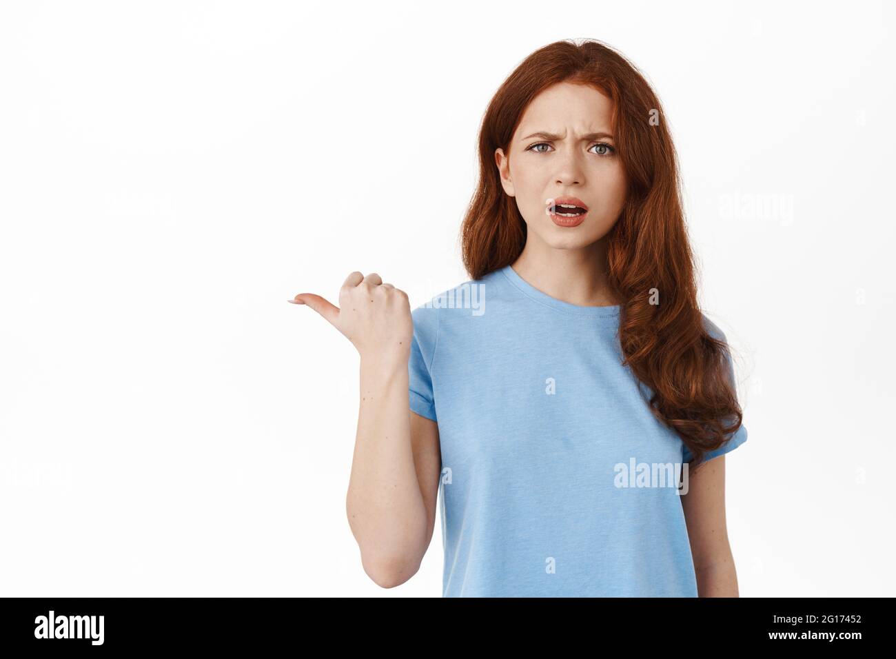 Confused and annoyed redhead girl pointing finger left, showing something strange and frustrating, cant understand what is that aside, standing in t Stock Photo