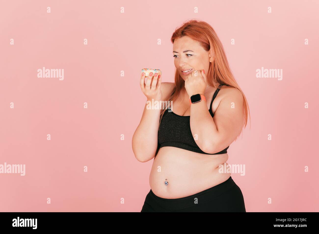 Oversized young woman wearing sports outfit looks at sweet donut junk food doubts but wants to eat dessert, standing on pink background, copy space. Stock Photo