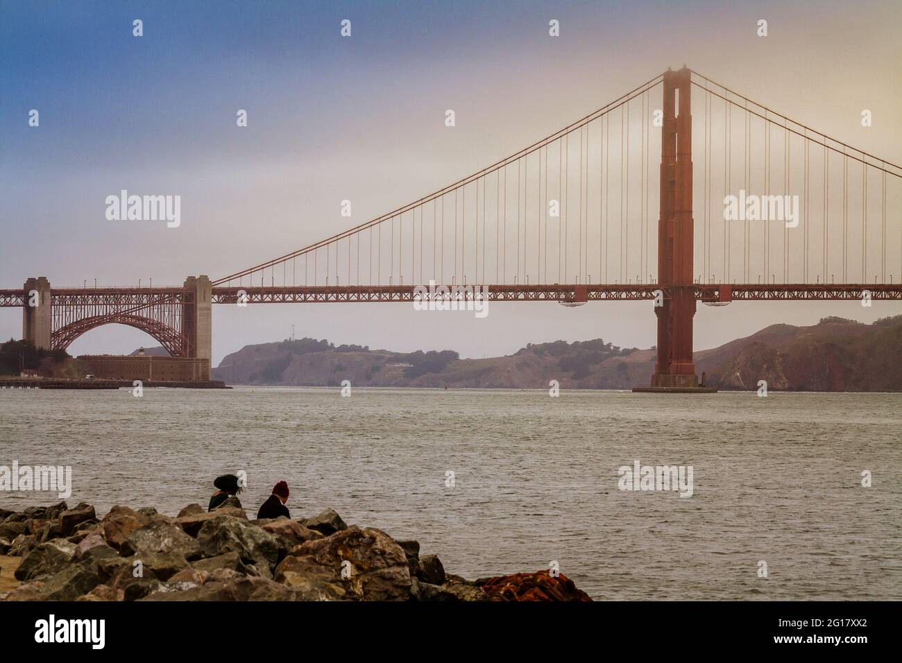 Golden Gate Bridge on a foggy day and two people sitting by San Francisco Bay Stock Photo