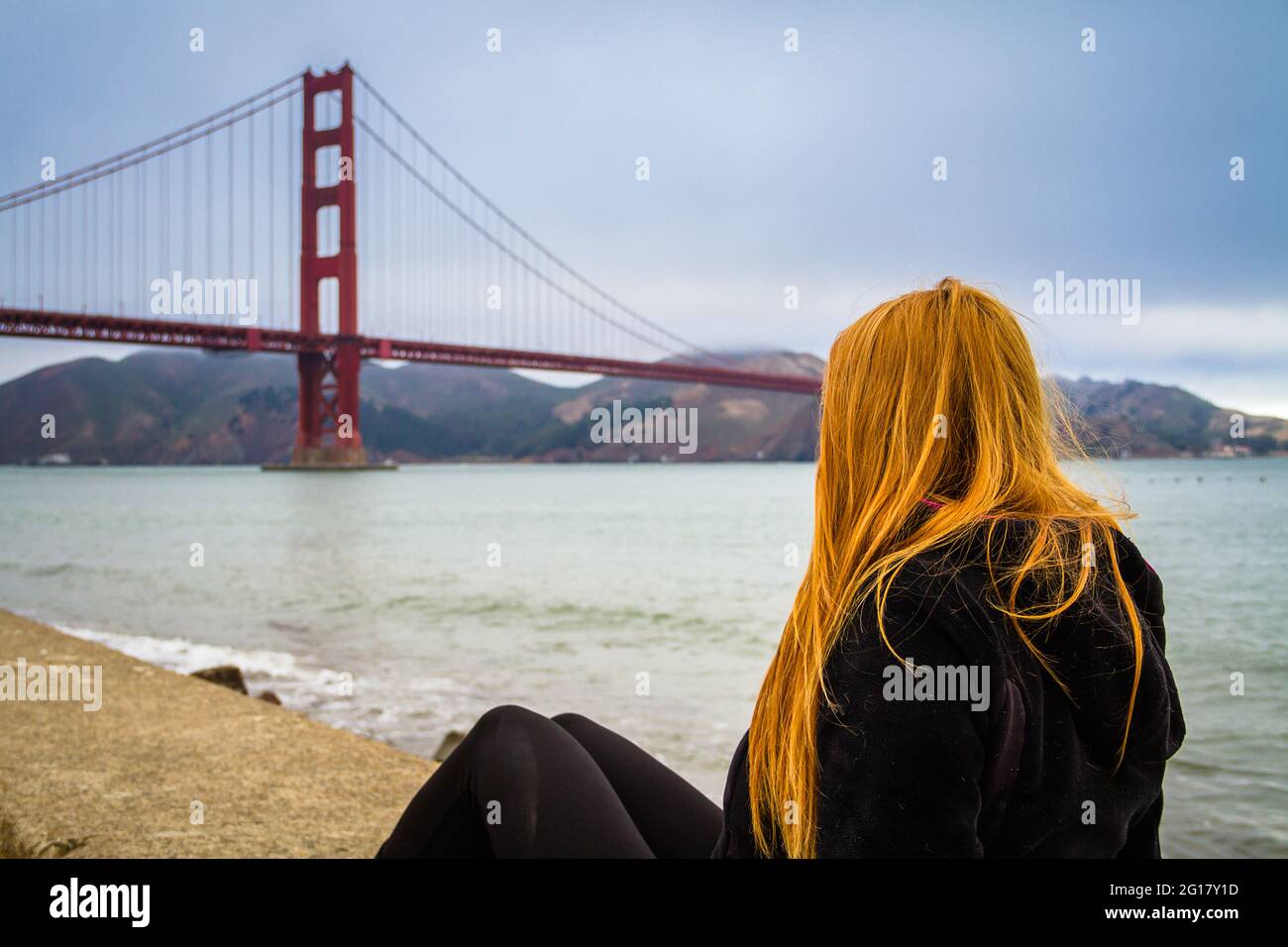 A young woman with red long hair sitting by San Francisco Bay watching the Golden Gate Bridge Stock Photo