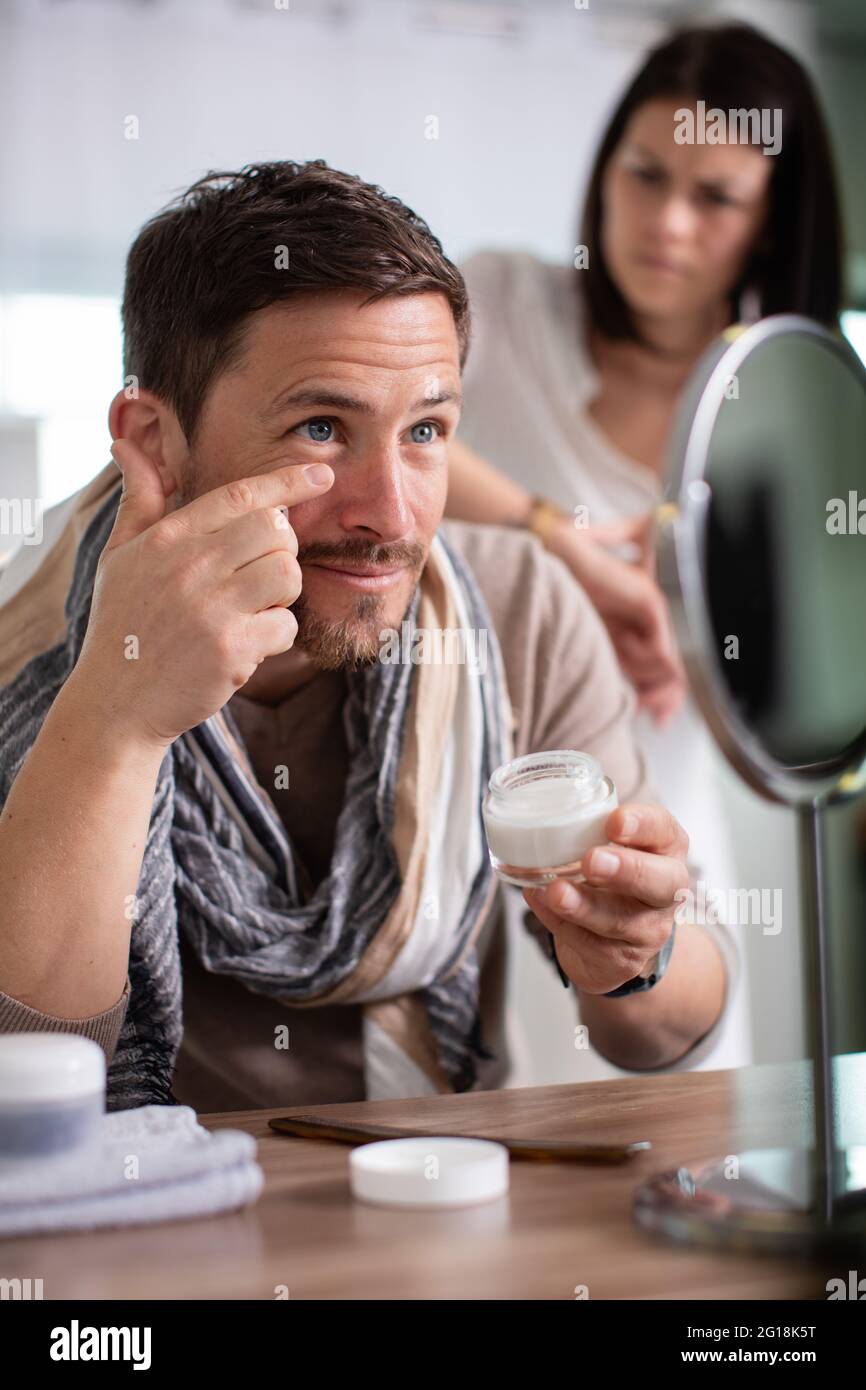 man applys moisturiser while impatient woman waits to go out Stock Photo