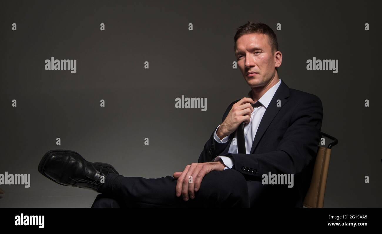 Portrait of a pensive confident man, in a business suit, sitting cross-legged against a dark background. Stock Photo