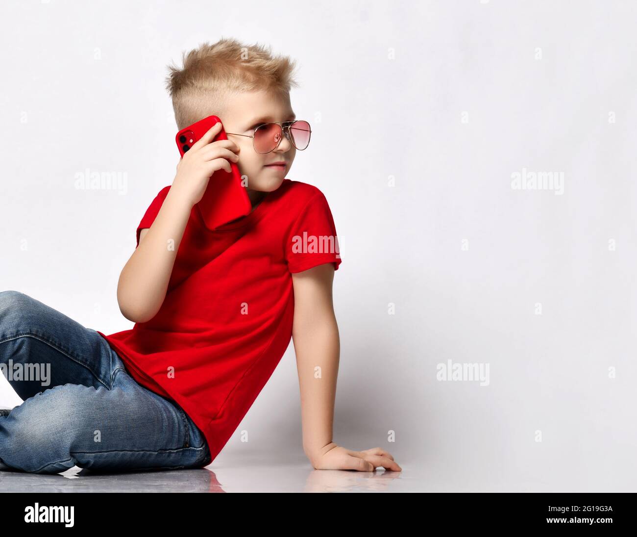 Stylish kid boy child in red t-shirt, jeans, sneakers and sunglasses sitting on floor talking on smartphone looking away Stock Photo