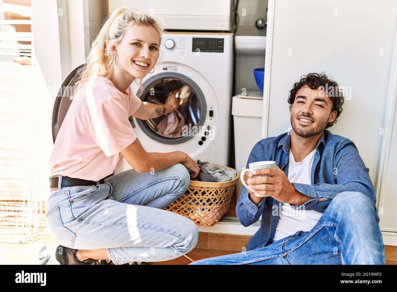 Young couple smiling happy drinking coffee while doing laundry at home ...