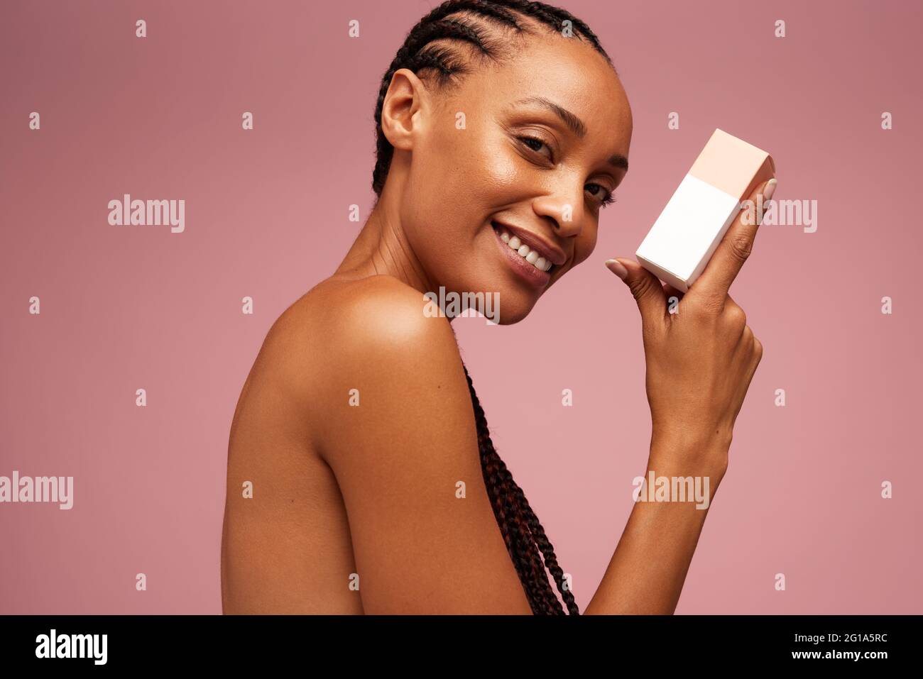 Attractive female model holding beauty product against pink background. Beautiful woman with cosmetic product looking at camera. Stock Photo