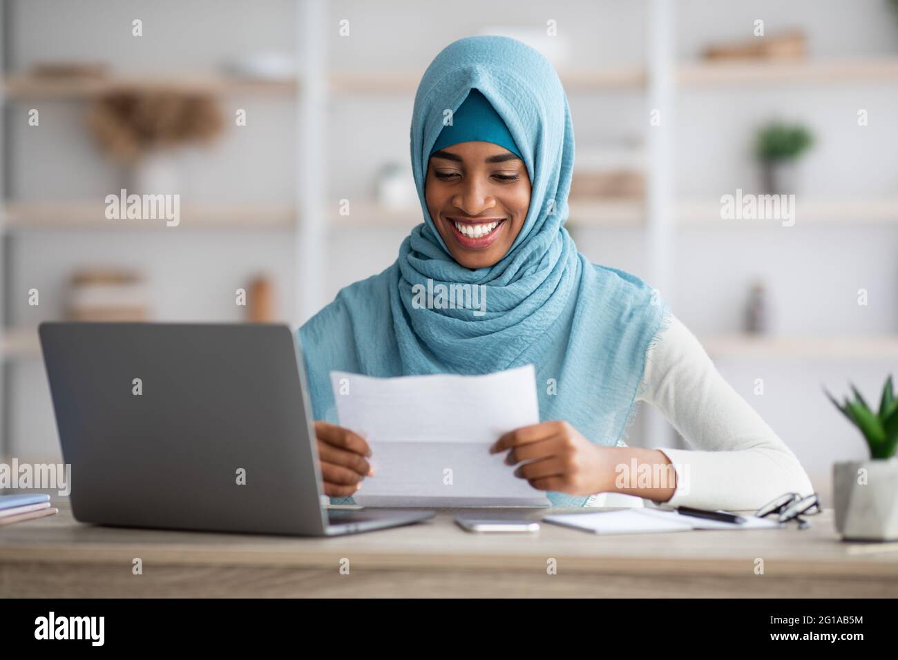 Happy Black Muslim Woman In Hijab Sitting At Desk And Reading Letter ...