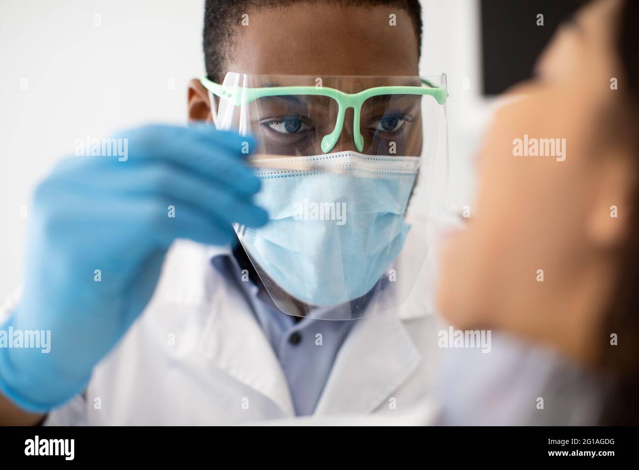 Black Male Dentist In Protective Mask And Face Shield Examining Patient's Teeth Stock Photo