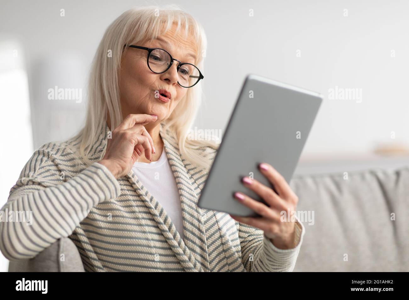 Surprised mature woman using tablet sitting on couch Stock Photo