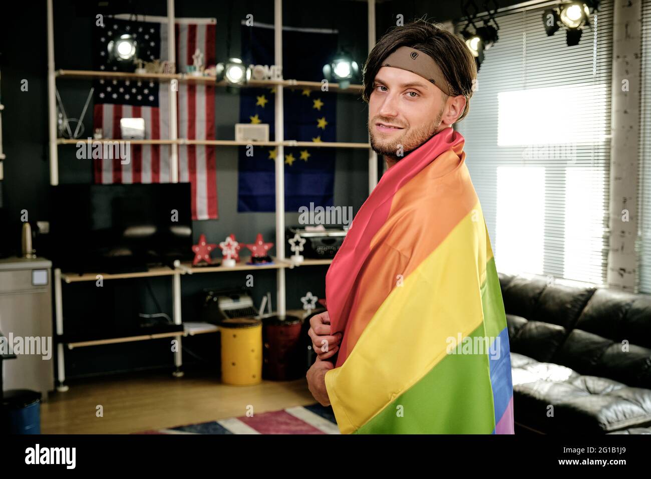 Young gay man with pride flag standing in living-room and looking at camera Stock Photo