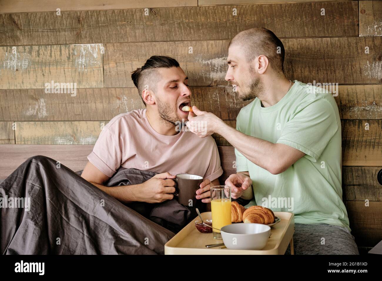 Young gay man putting cookie into mouth of his boyfriend during breakfast in bed Stock Photo