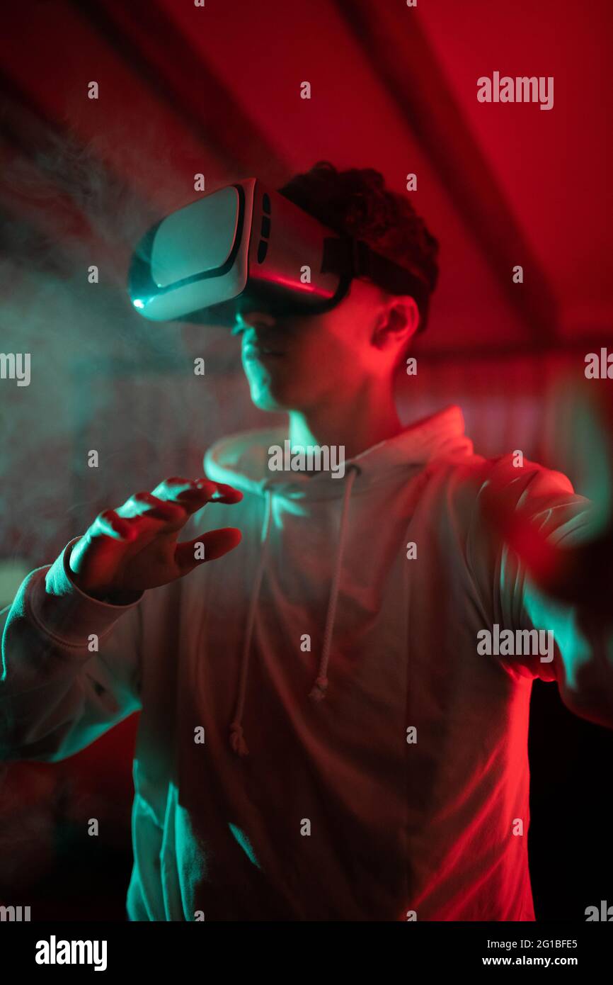 man wearing hoodie while using VR goggles and standing in studio with red neon lights Stock Photo
