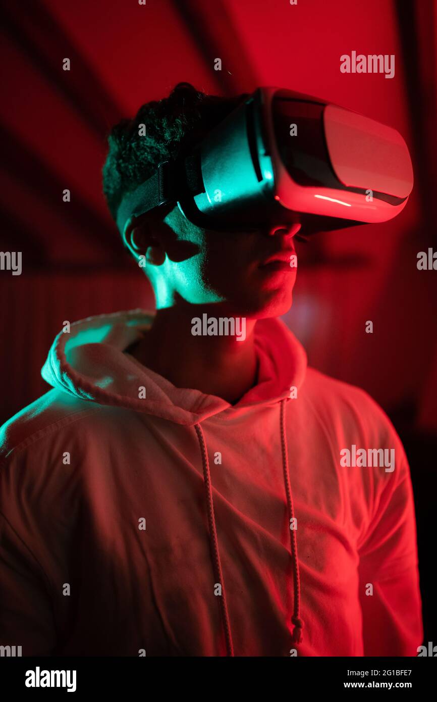 man wearing hoodie while using VR goggles and standing in studio with red neon lights Stock Photo