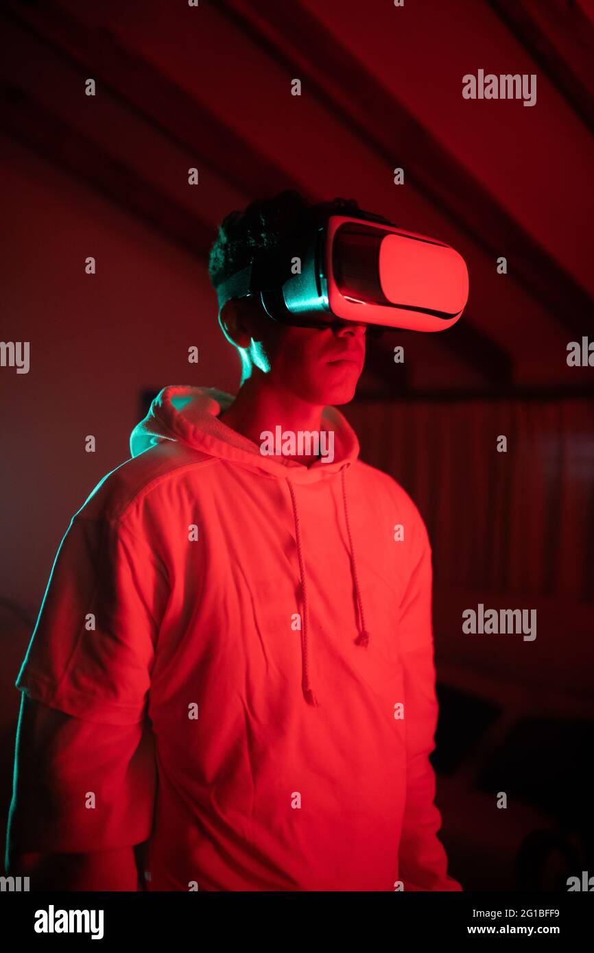 man wearing hoodie while using VR goggles and standing in studio with red neon lights Stock Photo