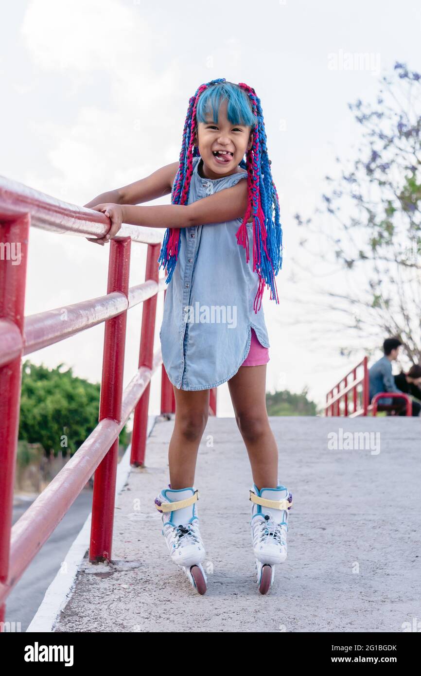 Cheerful ethnic child with tongue out and colorful braids roller skating while looking at camera in town Stock Photo